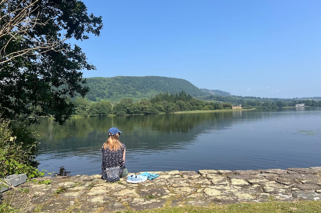 A woman enjoying the view at Lake of Menteith, her partners clothes are beside her like he's jumped in the water